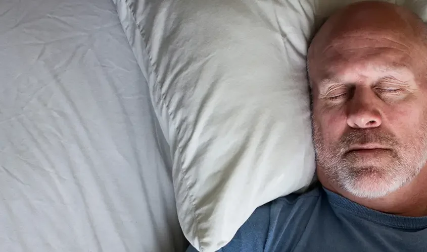 A man is sleeping soundly in a bed, with a blanket draped over him in a calm and serene bedroom environment.