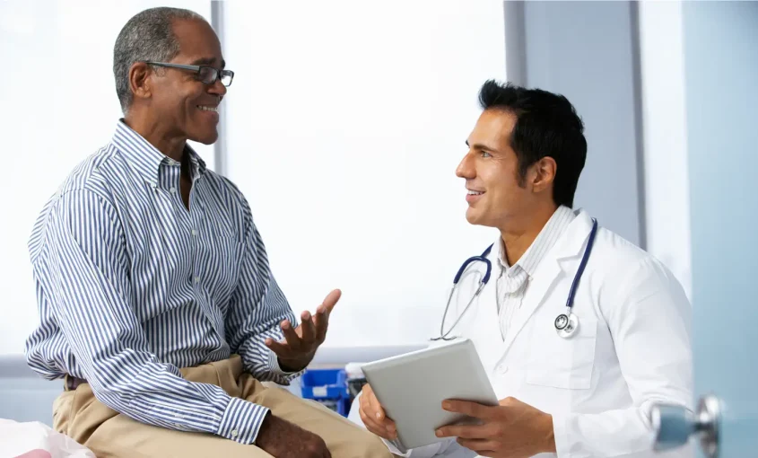 A doctor in a white coat converses with a patient in a medical office, discussing health concerns and treatment options.