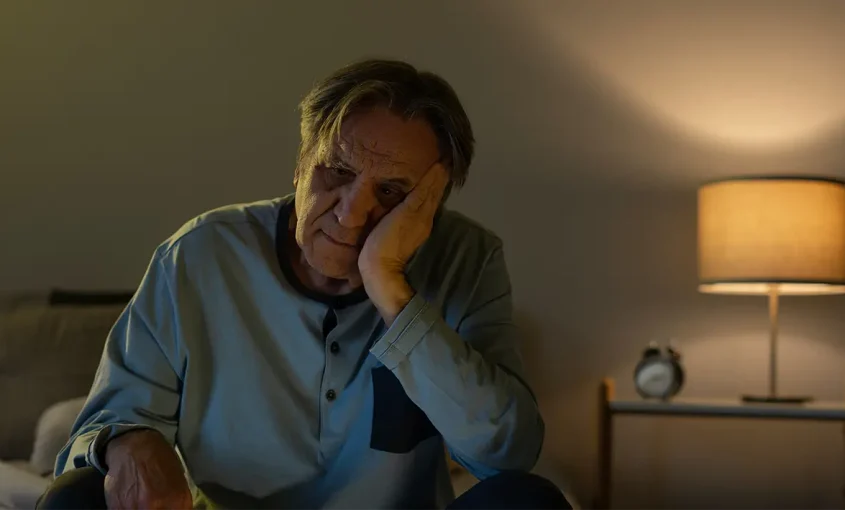 A man sitting on a bed in a dimly lit room, struggling to get to sleep.
