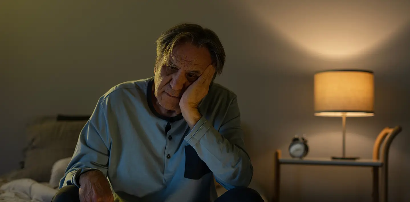 A man sitting on a bed in a dimly lit room, struggling to get to sleep.